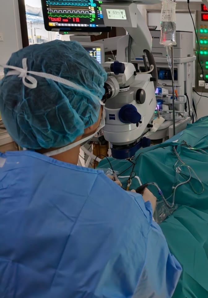 Dr Wong operating at the surgical microscope in theatre, the patient fully draped, with the anaesthetic monitor behind.